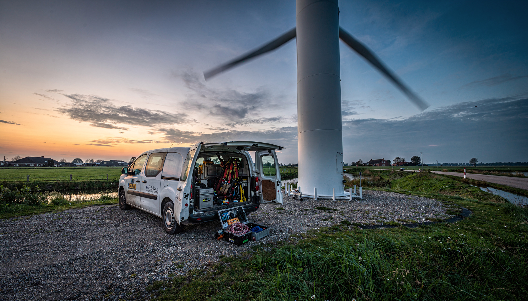 Onderhoudsmateriaal bij de voet van een windturbine in een Nederlandse polder tijdens de avondschemering.